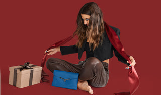 Botkier holiday gift guide main image model sitting with gift box and valentina flat crossbody in sapphire